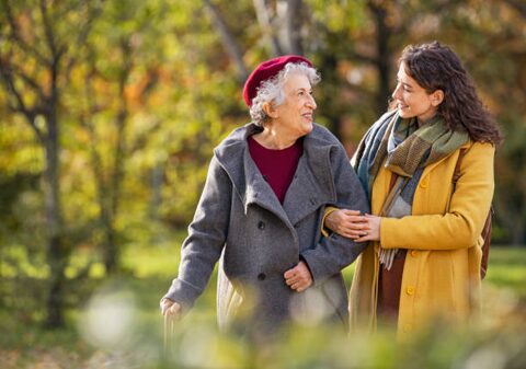 Two women walk together in a sunny autumn park, one elderly with a red hat and gray coat supported by a younger woman in a mustard coat.