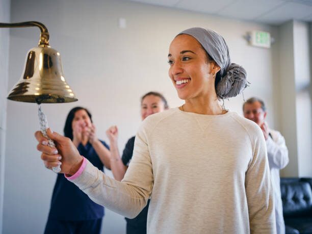 image8 Woman in a light sweater and gray head wrap rings a brass bell as colleagues cheer and applaud in a bright office room.
