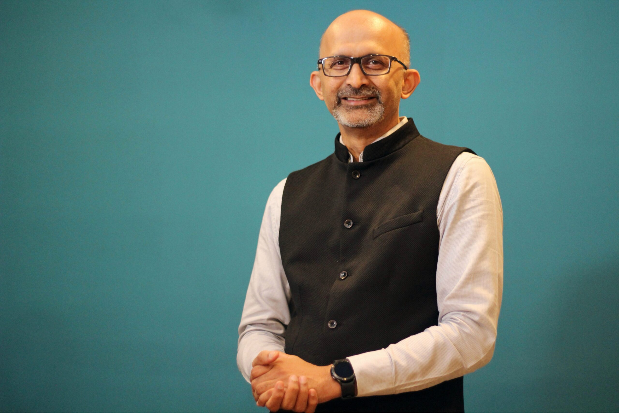 Man with glasses in a black waistcoat over a light shirt, standing against a teal wall with hands clasped.