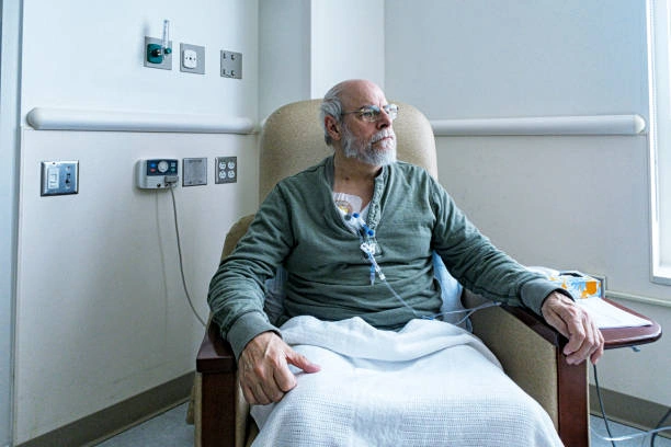 Older man with glasses sits in a hospital chair, connected to an IV and monitor cables.