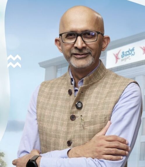 Man with glasses in a black waistcoat over a light shirt, standing against a teal wall with hands clasped.