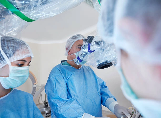 Surgeons in blue scrubs and masks perform operation under surgical microscope in a well-lit operating room
