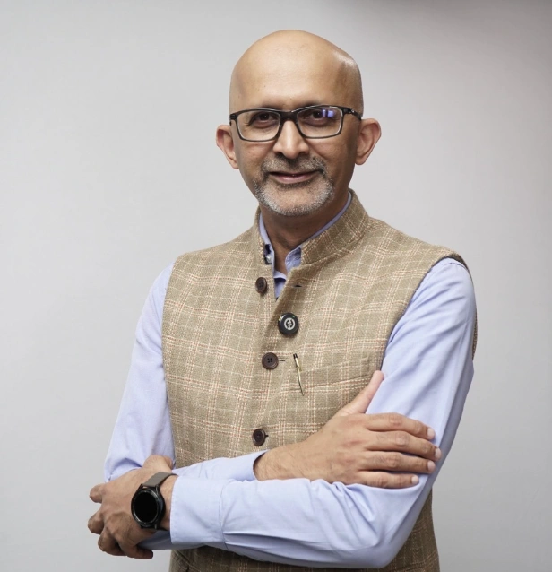Smiling bald man with glasses wearing a beige checkered vest over a light blue shirt, arms crossed.