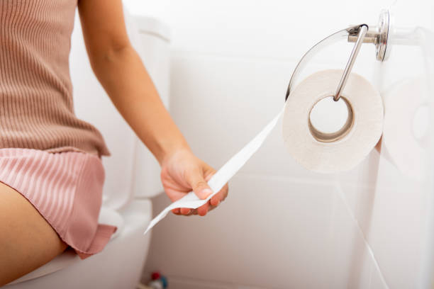 Person sitting on a toilet, pulling toilet paper from the wall-mounted roll holder.
