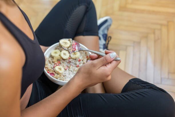 Person sitting on a wooden floor eating a bowl of oatmeal topped with banana slices.