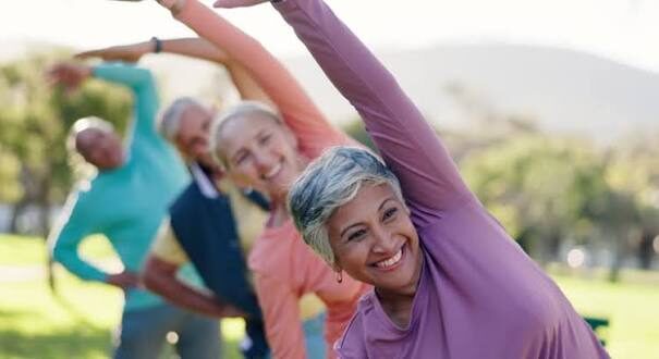 Senior adults doing a side-stretch yoga pose in a sunny park, smiling at the camera.