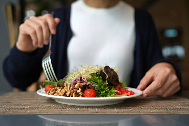 Person wearing a white top and dark cardigan forks a plate of salad with greens, cherry tomatoes, and walnuts at a table.