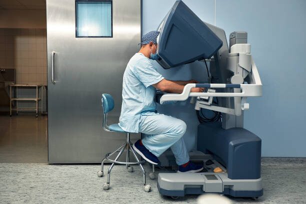 Medical professional in scrubs and a mask sits on a stool and operates a large diagnostic imaging machine in a hospital room, performing a scan.