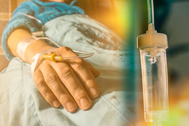 Close-up of a patient's hand with an IV line, resting on a hospital bed, holding a small yellow medical instrument; an IV drip is nearby.
